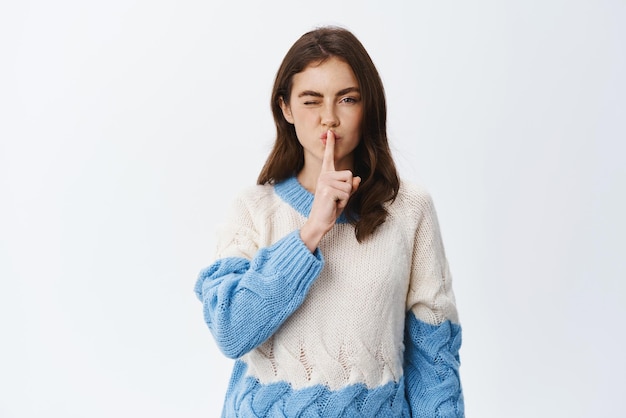 Jolie fille brune chut devant la caméra et clignant de l'œil en disant un secret et chut faisant signe chut debout en pull sur fond blanc