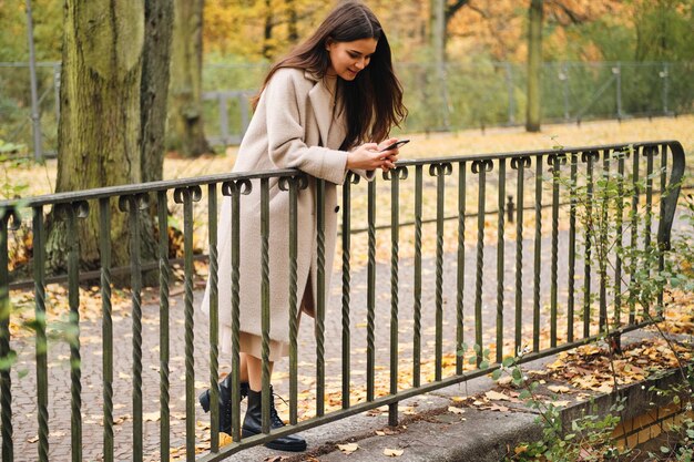 Jolie fille brune décontractée en manteau utilisant joyeusement un téléphone portable se reposant dans le parc d'automne seul