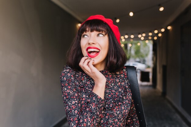 Jolie fille brune portant des vêtements de style français touchant son visage, se souvenait de quelque chose de drôle. Portrait d'adorable jeune femme aux cheveux noirs courts en béret rouge et costume vintage s'amusant à l'intérieur