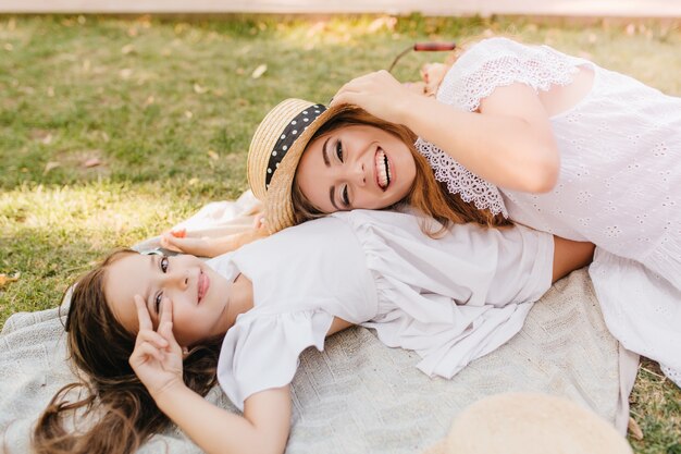 Jolie fille brune posant avec signe de paix sur l'herbe tandis que sa charmante mère tenant un chapeau de paille et riant. Jolie jeune femme en tenue de dentelle s'amuser avec sa fille sur une couverture dans le parc.