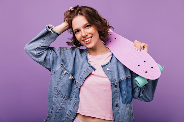 Jolie fille brune posant en veste en jean. Winsome jeune femme debout avec planche à roulettes.