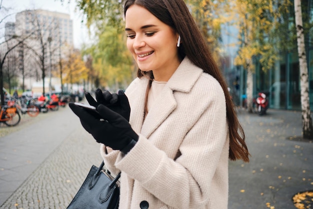 Photo gratuite jolie fille brune souriante en manteau utilisant joyeusement un téléphone portable se promenant seule dans la rue de la ville