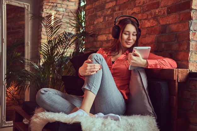 Une jolie fille brune souriante vêtue de vêtements décontractés à l'aide d'une tablette tout en écoutant de la musique passionnante via de bons écouteurs. Détente dans une chambre à l'intérieur loft.