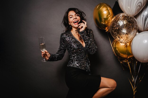 Jolie fille brune avec un verre à vin dansant sur fond noir Modèle féminin élégant profitant d'une fête d'anniversaire