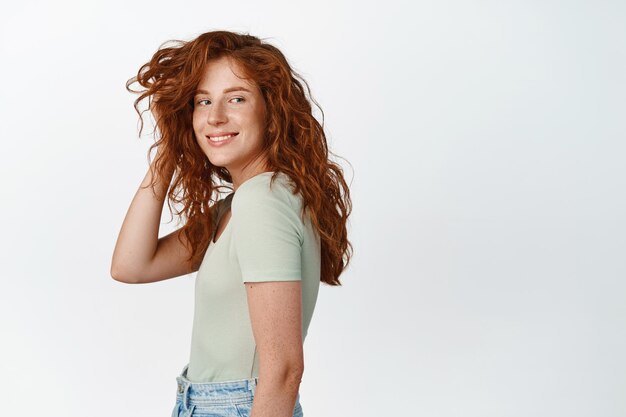 Jolie fille candide aux cheveux bouclés rouges et aux taches de rousseur jouant avec la coupe de cheveux et souriante heureuse à la caméra debout en t-shirt sur fond blanc