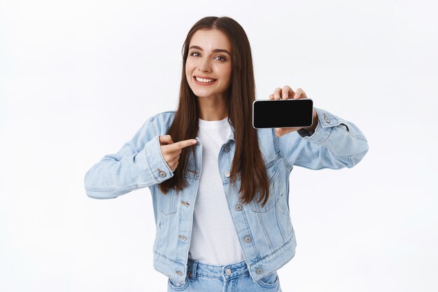 Jolie fille caucasienne moderne sortante en veste en jean, tenant le smartphone horizontalement, montrant et pointant l'écran mobile, souriant heureux, donne des conseils sur l'application de téléchargement, fond blanc