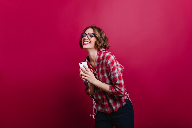 Jolie fille en chemise à carreaux riant les yeux fermés sur le mur bordeaux. Photo intérieure d'une femme aux cheveux courts bouclés émotionnelle dans des verres tenant le téléphone.