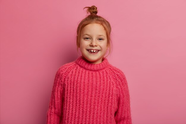 Jolie fille de cinq ans pose, exprime des émotions positives, a les cheveux roux, porte un pull d'hiver chaud, heureuse d'être photographiée, pose contre un mur rose. Émotions sincères et enfants.
