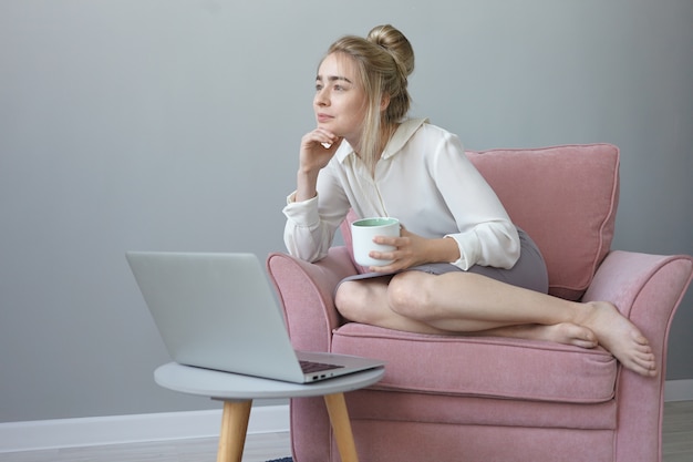 Jolie fille avec une coiffure en désordre ayant une expression faciale de rêve, prenant un café, assise dans un fauteuil confortable devant un ordinateur portable ouvert, pensant aux projets futurs et aux vacances d'été