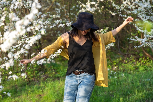 Photo gratuite une jolie fille dans un chapeau parmi les arbres en fleurs apprécie l'odeur des fleurs printanières.