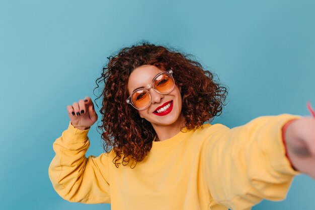 Jolie fille dans des lunettes de soleil orange vif et avec des lèvres brillantes posant sur l'espace bleu. Femme en haut jaune prend selfie.