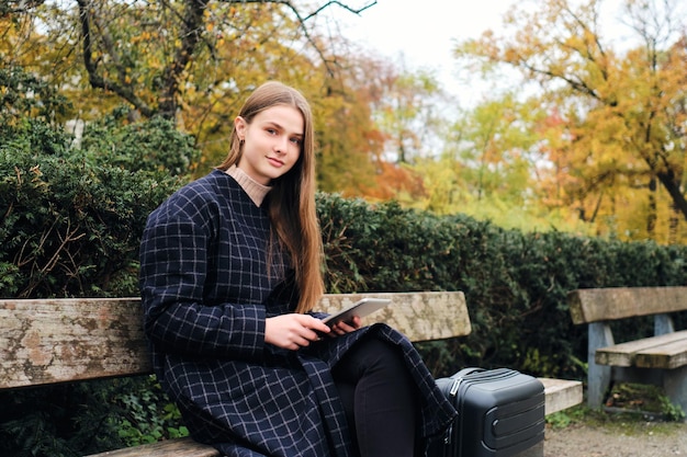 Jolie fille décontractée à l'aide d'une tablette assise avec une valise sur un banc dans le parc de la ville