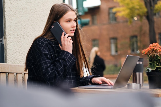 Photo gratuite jolie fille décontractée parlant sur un téléphone portable et surfant sur internet sur un ordinateur portable dans un café de rue