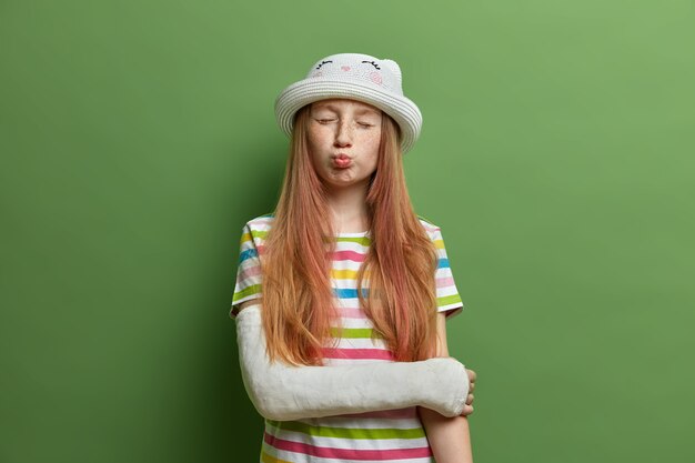 Une jolie fille drôle fait la grimace et fait la bouche aux lèvres, a le visage taché de rousseur et de longs cheveux foxy, pose avec un plâtre sur un bras cassé, s'est blessée pendant les vacances d'été, porte un t-shirt et un chapeau à rayures