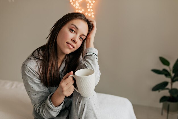 Jolie fille élégante en costume gris avec des cheveux noirs est réveillée et assise sur le lit avec une tasse de café dans une chambre élégante et lumineuse avec des lumières en arrière-plan