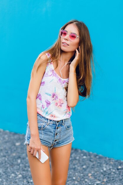 Jolie fille élégante sur fond bleu se dresse et écoute de la musique sur des écouteurs sur smartphone.