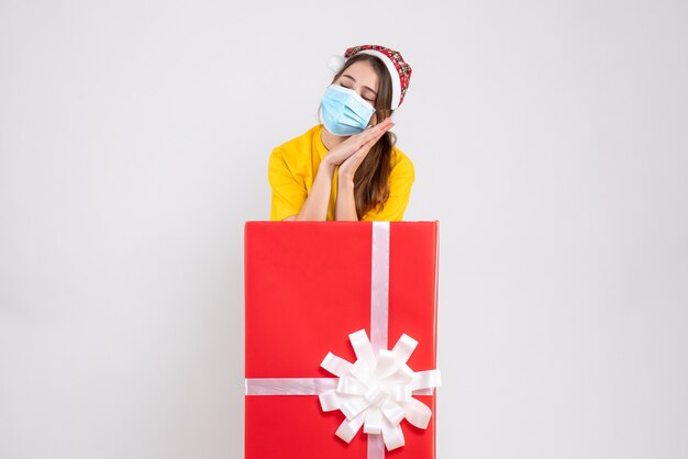 Jolie fille endormie avec bonnet de Noel debout derrière un gros cadeau de Noël sur blanc