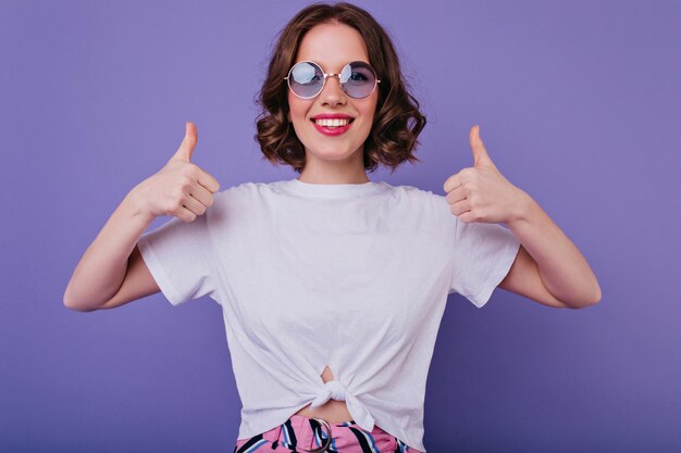 Jolie fille européenne dans des lunettes de soleil à la mode exprimant des émotions positives et posant avec les pouces vers le haut Photo en studio d'une dame blanche de bonne humeur debout sur fond violet