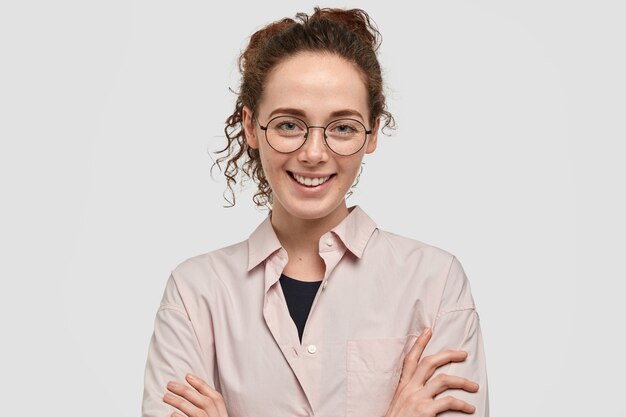 Jolie fille européenne avec un sourire charmant, garde les bras croisés, porte des lunettes rondes