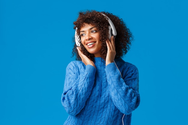 Jolie fille féminine afro-américaine sensuelle avec coupe de cheveux afro, portant un pull d'hiver, regardant à gauche avec un sourire agréable, portant des écouteurs, écoute des chansons.