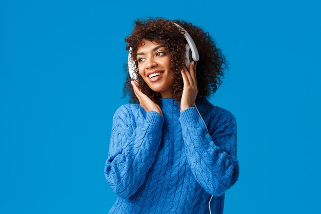 Jolie fille féminine afro-américaine sensuelle avec coupe de cheveux afro, portant un pull d'hiver, regardant à gauche avec un sourire agréable, portant des écouteurs, écoute des chansons.