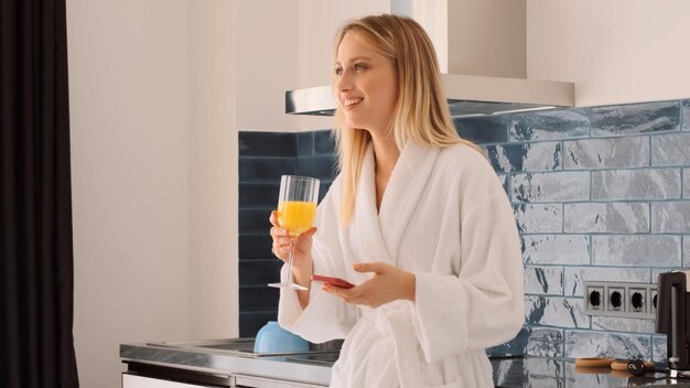 Jolie fille gaie en peignoir blanc buvant du jus d'orange avec un smartphone au petit-déjeuner dans la cuisine à la maison