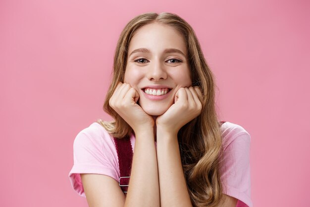 Jolie fille gentille féminine et tendre avec un tatouage et des cheveux ondulés penchés la tête sur les mains comme si elle écoutait une histoire amusante en regardant avec un large sourire et un regard d'admiration rêveur sur fond rose.