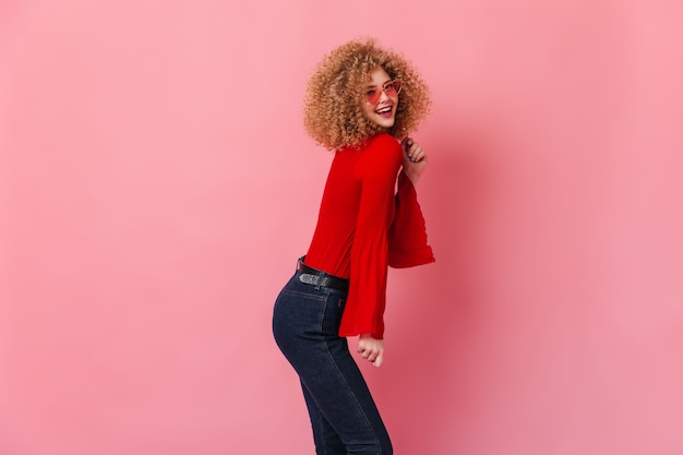 Photo gratuite jolie fille en haut rouge et jeans foncés rit. dame à lunettes de soleil élégantes posant sur l'espace rose.