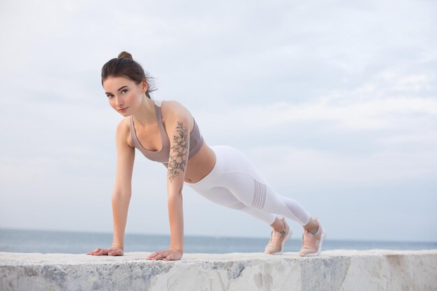 Jolie fille en haut sportif et leggings blancs pratiquant le yoga avec vue sur la mer en arrière-plan. Jeune femme regardant pensivement à huis clos tout en s'entraînant au bord de la mer