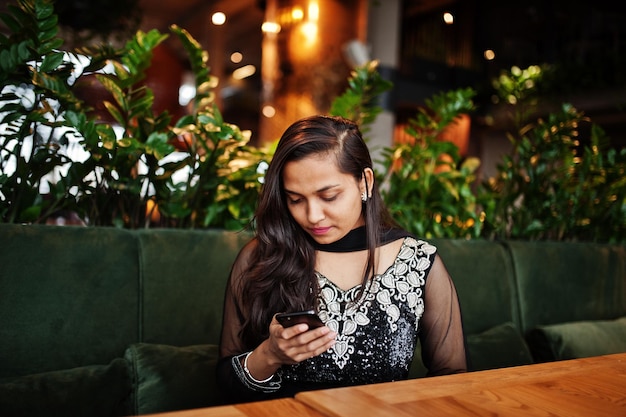 Photo gratuite jolie fille indienne en robe sari noire posée au restaurant avec un téléphone portable à portée de main