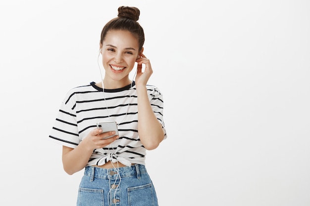 Jolie fille insouciante souriante, mettre des écouteurs pour écouter un podcast ou de la musique sur téléphone mobile