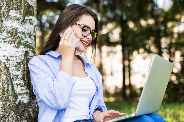 Jolie fille en jean bleu travaille avec un ordinateur portable dans le parc de la ville parlant avec téléphone