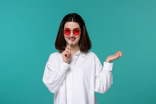 Jolie fille jeune jolie adorable fille brune en chemise blanche léchant une sucette à lunettes rouges