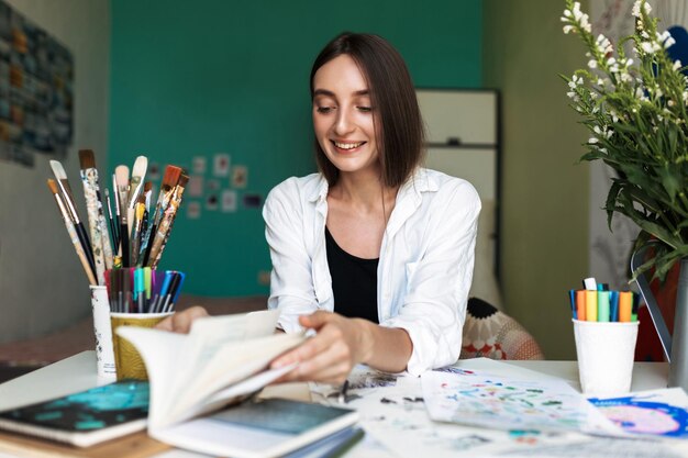Jolie fille joyeuse assise au bureau avec des peintures feuilletant joyeusement un livre tout en passant du temps à la maison