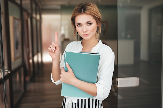 Jolie fille avec des livres à la main regardant à huis clos et ne montrant aucun geste. Portrait de jeune fille aux cheveux blonds en chemise blanche debout près de la porte et tenant des cahiers à la main.