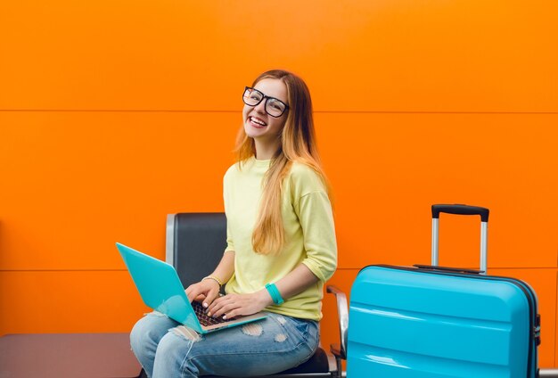 Jolie fille à lunettes noires aux cheveux longs est assise sur une chaise sur fond orange. Elle a un ordinateur portable bleu et une valise. Elle porte un pull jaune et sourit à la caméra.