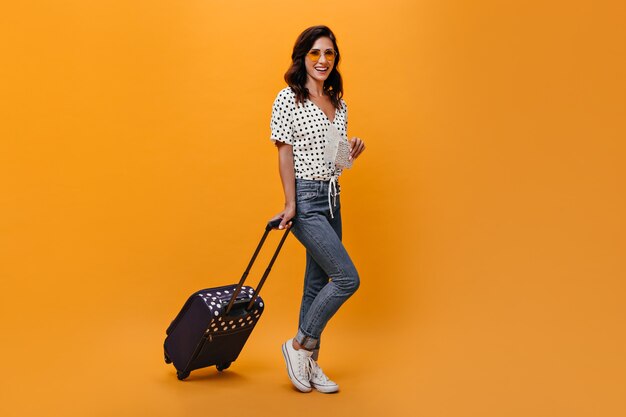 Jolie fille à lunettes porte valise sur fond orange. Brunette aux cheveux ondulés dans des lunettes de soleil en chemisier blanc à pois noirs posant.