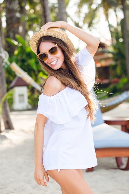 Jolie fille en lunettes de soleil marron et chapeau de paille sourit largement et tient son chapeau avec une main