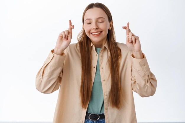 Une jolie fille pleine d'espoir ferme les yeux et croise les doigts pour la chance en priant en faisant un vœu en demandant de bons résultats en anticipant les nouvelles debout sur fond blanc