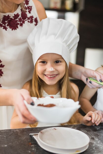 Jolie fille portant la toque debout devant la mère pendant la cuisson dans la cuisine