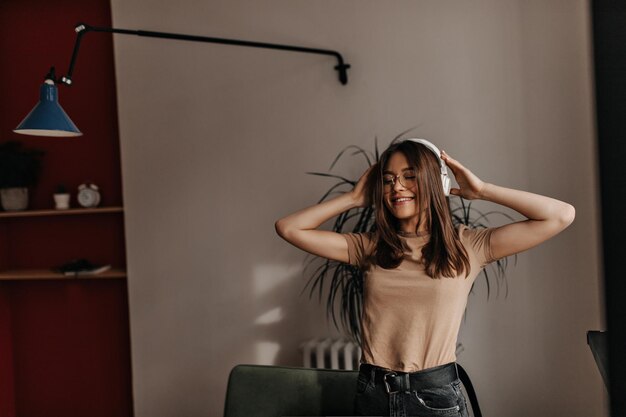 Jolie fille porte des écouteurs blancs Photo d'une femme en jeans noirs et t-shirt posant les yeux fermés dans une pièce lumineuse