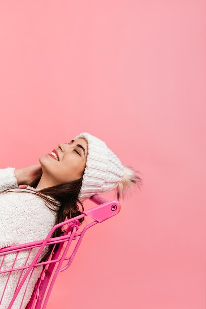 Jolie fille en riant, posant dans un chariot de supermarché rose. Plan d'une brune en vêtements d'hiver blancs sur un mur isolé.