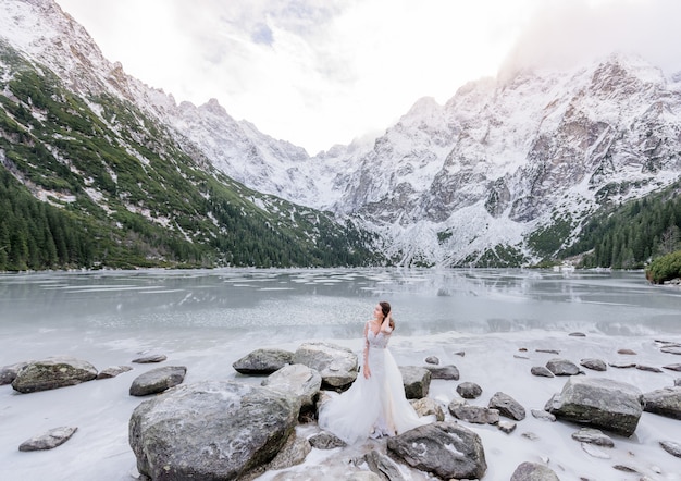 Jolie fille en robe blanche se tient devant un lac gelé entouré de montagnes enneigées