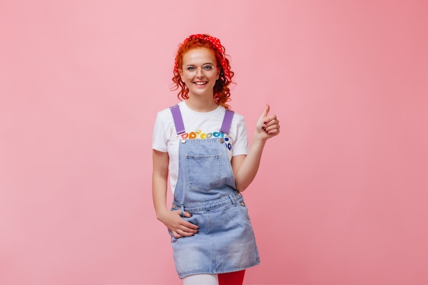 Jolie fille en robe d'été en jean et t-shirt blanc avec un sourire regardant la caméra et montrant les pouces vers le haut sur un mur isolé