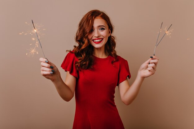 Jolie fille en robe rouge tenant des cierges magiques à la fête du nouvel an. Femme élégante au gingembre posant avec des lumières du Bengale à Noël.