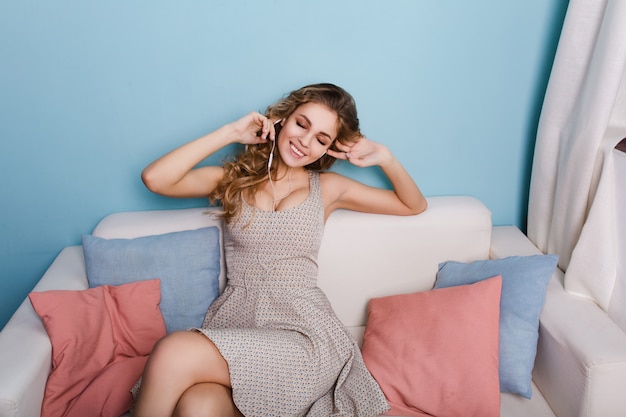 Jolie fille sexy aux cheveux bouclés blonds est assise sur un canapé en studio avec des murs bleus et des oreillers colorés.