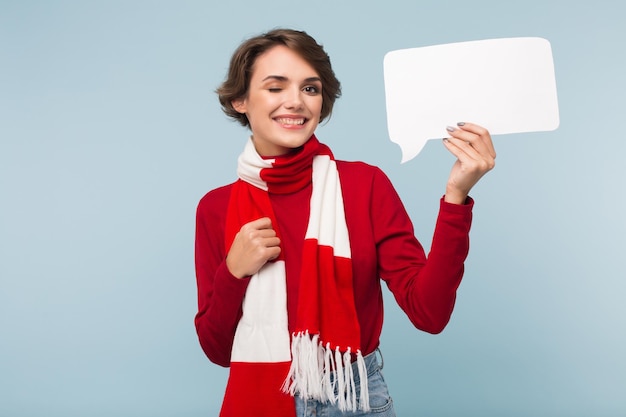 Jolie fille souriante aux cheveux courts foncés en pull rouge et écharpe clignant joyeusement de l'œil tout en regardant à huis clos avec une forme de papier blanc de message à la main sur fond bleu