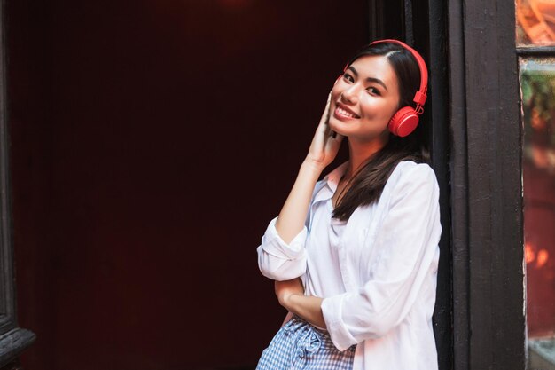 Jolie fille souriante en chemise blanche regardant joyeusement à huis clos s'appuyant sur la vieille porte tout en écoutant de la musique dans des écouteurs rouges