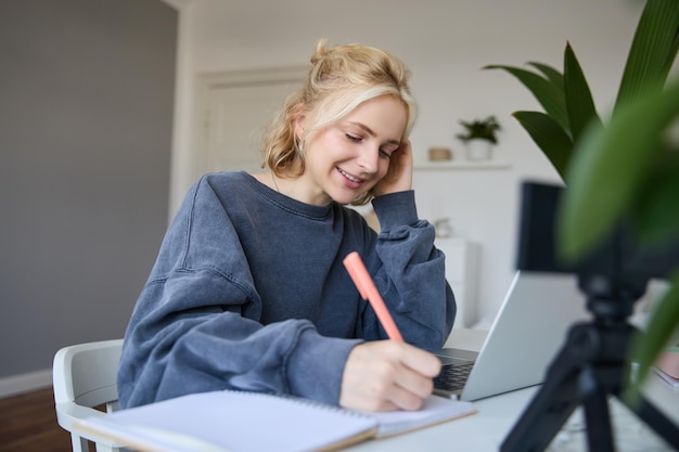 Photo gratuite une jolie fille souriante est assise dans une pièce, écrit des notes, fait des devoirs, enregistre une vidéo d'elle-même.