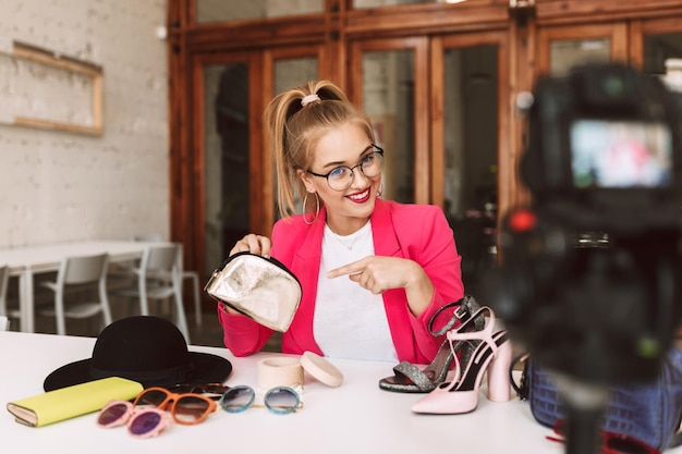 Jolie fille souriante à lunettes et veste rose enregistrant joyeusement une nouvelle vidéo de mode pour son vlog